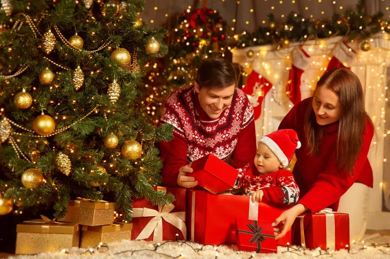 A mom and dad and baby sitting under a Christmas tree.